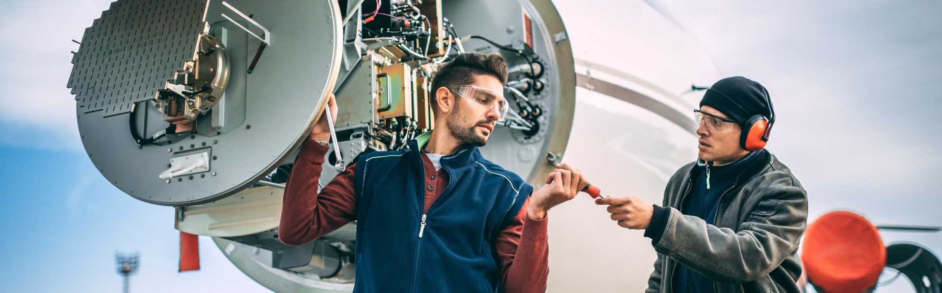 Two aircraft mechanics working on a radar array under the opened up nose cone of a private airplane parked on an airport field